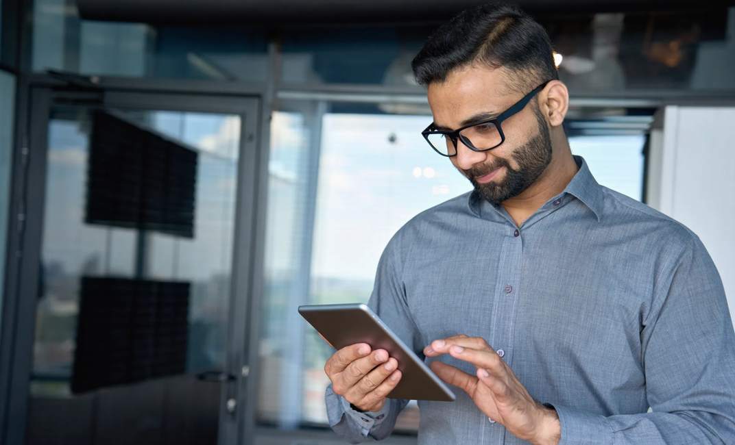 Young indian marketing business manager expert in glasses standing in modern corporate office using digital tablet device searching analyzing project results. Business technologies concept.